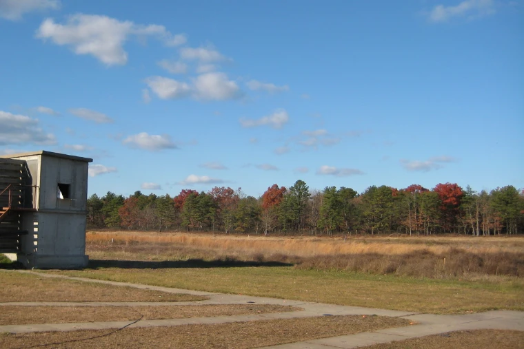 A concrete skeet house at the edge of a shooting field, with pines and red autumn oaks beyond under a blue sky (2007).