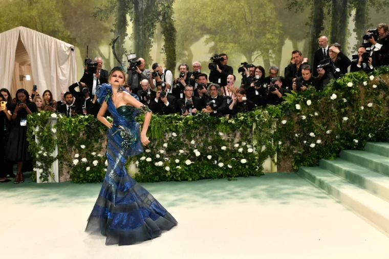 A guest in a blue and green iridescent gown with a feathered headpiece before the painted forest and white roses, in 2024. The Costume Institute Benefit at the Metropolitan Museum, New York, in the season calendar of The Mansa Court
