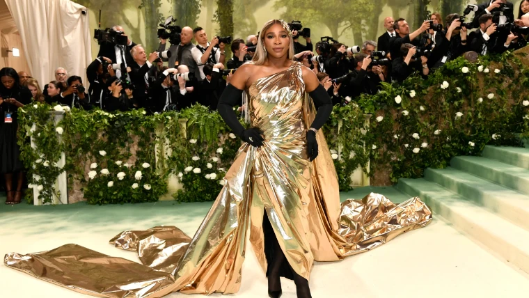 A guest in a draped gold lamé gown with black opera gloves before the garden wall of white roses, in 2024. The Costume Institute Benefit at the Metropolitan Museum, New York, in the season calendar of The Mansa Court
