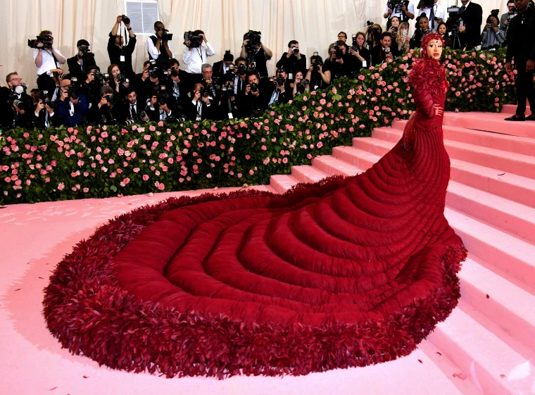 A guest in a red quilted gown with a vast feathered train on the pink-carpeted steps, before a wall of roses and photographers, in 2019. The Costume Institute Benefit at the Metropolitan Museum, New York, in the season calendar of The Mansa Court