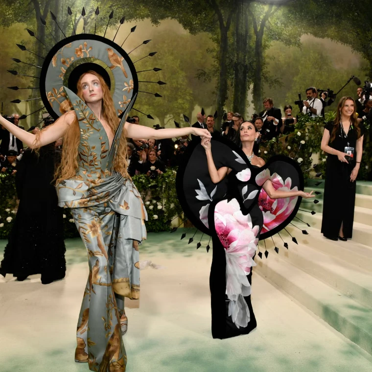 Two guests in sculptural couture, a painted silk suit beneath a sunburst halo and a black gown with an oversized peony, on the garden steps in 2024. The Costume Institute Benefit at the Metropolitan Museum, New York, in the season calendar of The Mansa Court