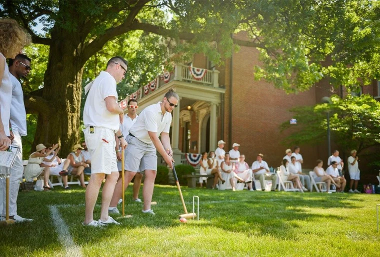 Players in whites at croquet on a shaded lawn before guests seated beneath the trees, at The Croquet and Lawn Party in New York, an afternoon in the season calendar of The Mansa Court