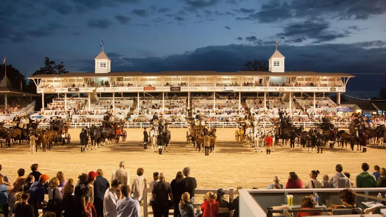 Four-in-hand coaches drawn up before the lit grandstand at dusk. The Devon Horse Show and Country Fair at the Devon Show Grounds, Pennsylvania, in the season calendar of The Mansa Court