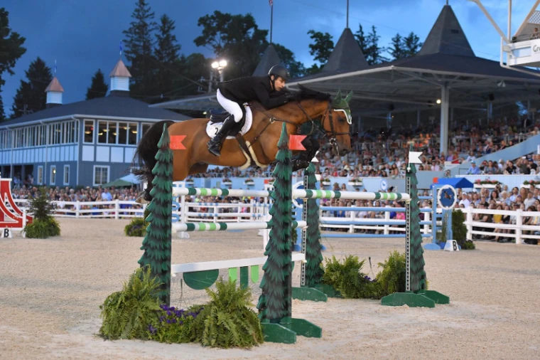 A bay clears a green fence in the Grand Prix before a full grandstand at evening. The Devon Horse Show and Country Fair at the Devon Show Grounds, Pennsylvania, in the season calendar of The Mansa Court