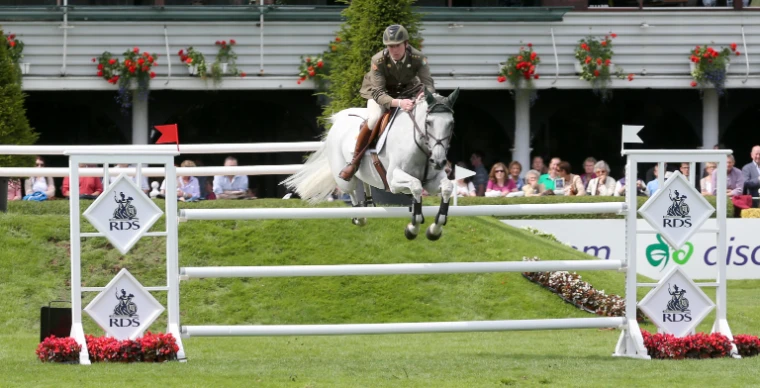 A rider in Irish Army uniform jumps a grey horse over a white fence in the main arena, with flower boxes and spectators along the stand behind (2012).