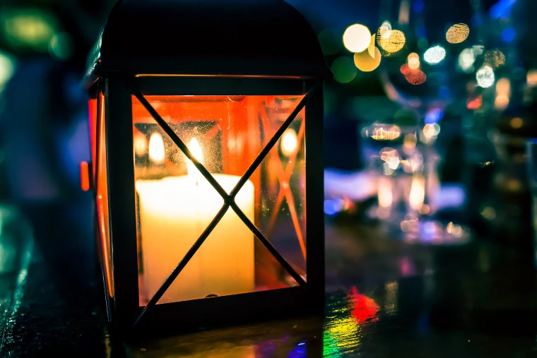 A candle burning inside a small black lantern on a dark table at night, with wine glasses blurred into coloured points of light behind (2014).