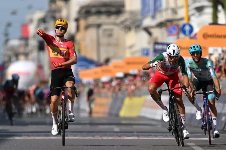 A rider punches the air as he takes a stage in a sprint finish on a city boulevard, his rivals at his shoulder. The Giro d’Italia, in the season calendar of The Mansa Court