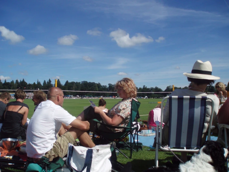 Spectators relax on picnic chairs and rugs at the edge of the polo field, one in a panama hat, under a blue sky with scattered clouds (2007).