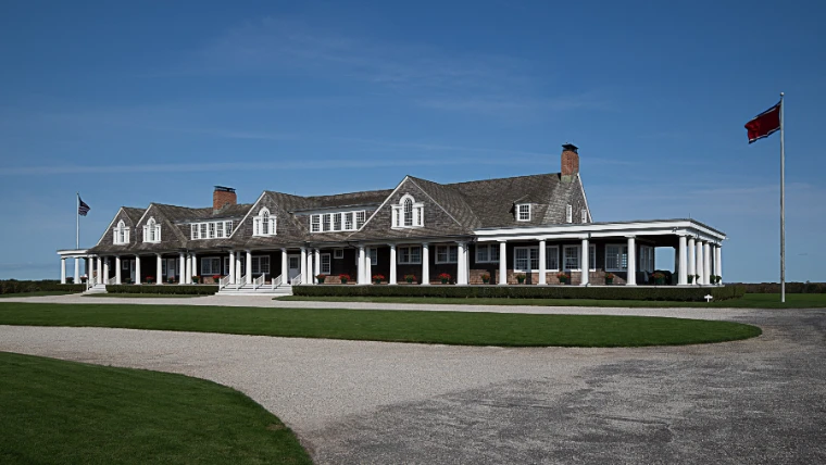 A shingled clubhouse with a white-columned veranda and a flag on a tall pole stands beyond a curving gravel drive and lawn under a deep blue sky (2021).