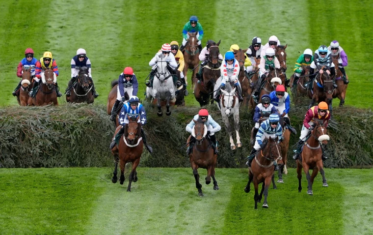 The field clearing a spruce-dressed fence on the Grand National course at Aintree, seen head-on across the striped turf, an April engagement in the season calendar of The Mansa Court