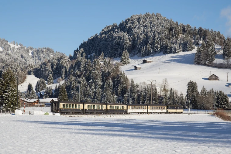 A cream and blue GoldenPass Classic train runs through a snow-covered valley below wooded hills (2015).