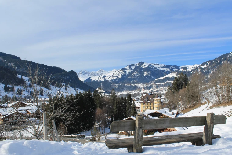 A wooden bench in the snow looking over the chalets of Gstaad to the snow-covered mountains beyond (2013).