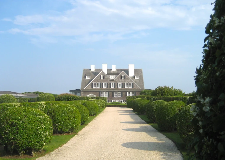 A large grey-shingled house with tall white chimneys stands at the end of a pale gravel drive lined with clipped round hedges under a summer sky (2010).