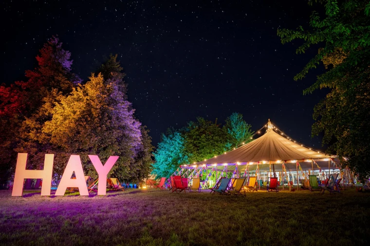 The great HAY letters and a lit marquee among coloured trees under a starry sky. The Hay Festival at Hay-on-Wye, on the Welsh border, in the season calendar of The Mansa Court
