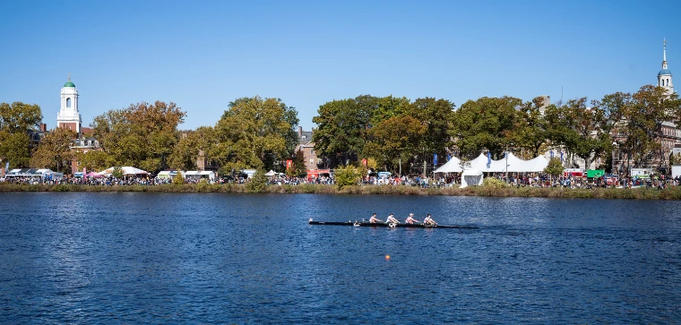 A crew rows past a far bank crowded with spectators and white tents under autumn trees and a white cupola tower (2022).