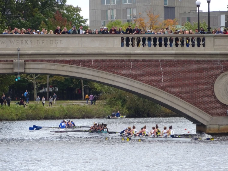 Rowing crews pass under the brick arch of the Weeks Footbridge as spectators line its balustrade above (2018).