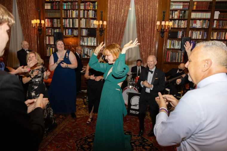 Guests dance on an oriental rug in a book-lined library as a band in black tie plays behind them (2025).