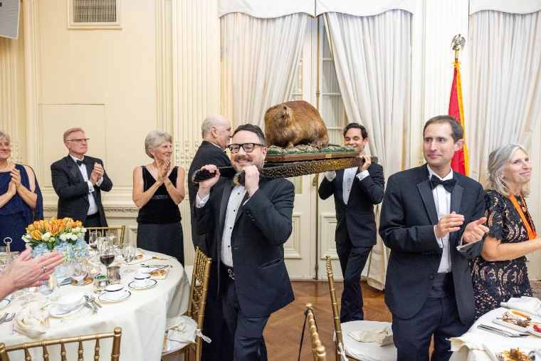 A man in black tie carries the Holland Society’s mounted beaver on his shoulder past the dinner tables as guests in evening dress applaud (2025).