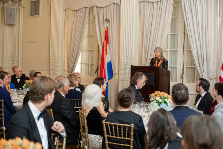 Louise Mirrer, wearing the Society’s medal on an orange ribbon, speaks from a lectern beside the Dutch flag to guests seated at dinner (2025).