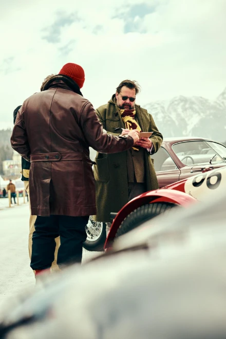 Two entrants in winter coats talk beside a red classic racing car on the lake. The I.C.E. St. Moritz, Lake St. Moritz, St. Moritz, Switzerland, February 2023, in the season calendar of The Mansa Court