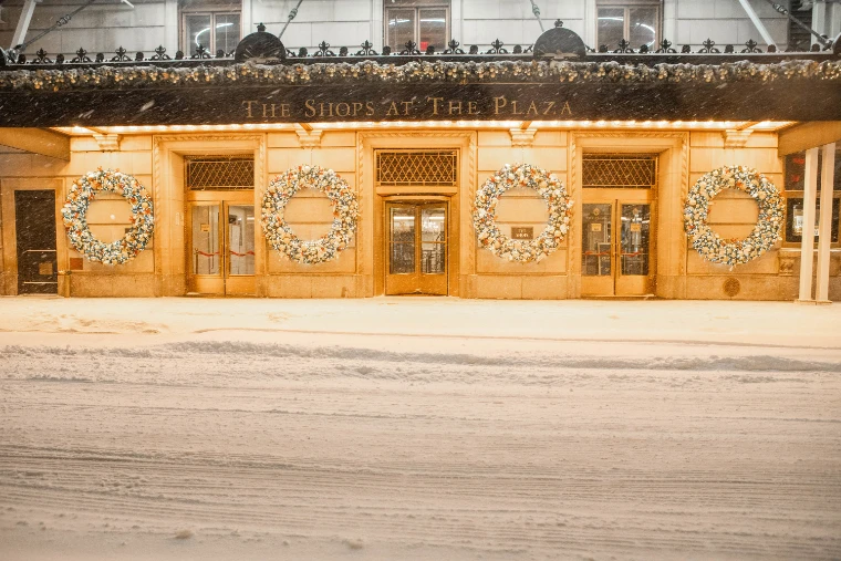 White Christmas wreaths on the gold-lit doors of the entrance to the Shops at the Plaza, beneath its canopy, the street in front deep in fresh snow at night (2020).