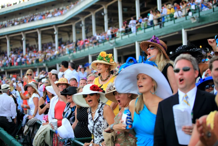Spectators in wide-brimmed Derby hats and summer dresses cheer at the rail below the grandstand. The Kentucky Derby at Churchill Downs, Louisville, in the season calendar of The Mansa Court