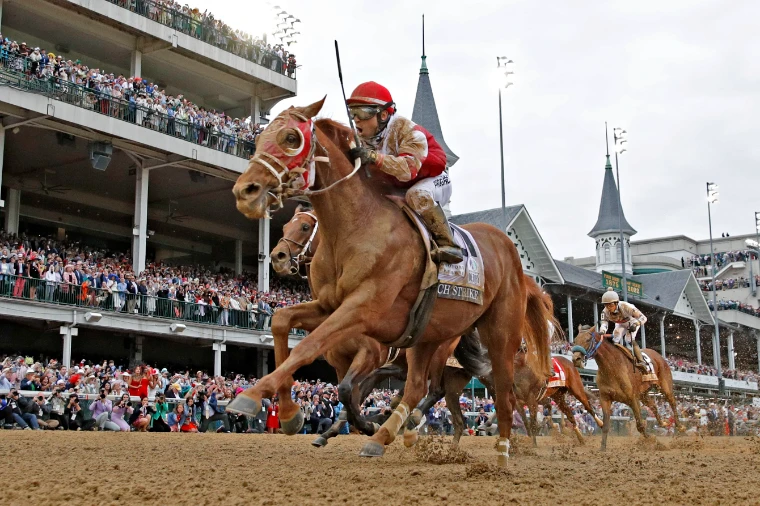 A chestnut and his jockey in red silks come down the stretch beneath the Twin Spires and a full grandstand, in 2022. The Kentucky Derby at Churchill Downs, Louisville, in the season calendar of The Mansa Court