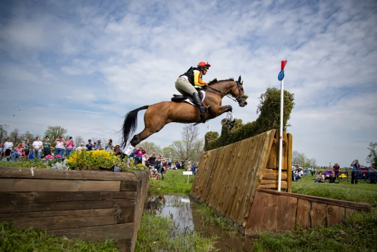A bay horse and rider in red and yellow flying a brush fence over the ditch on the cross-country course at the Kentucky Three-Day Event, a late-April engagement in the season calendar of The Mansa Court