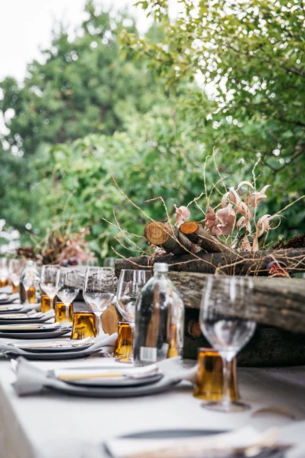 A long table laid with glasses, bottles and flowers beneath greenery, for The Long Table Sunday Lunches at the Court in New York, June Sundays in the season calendar of The Mansa Court