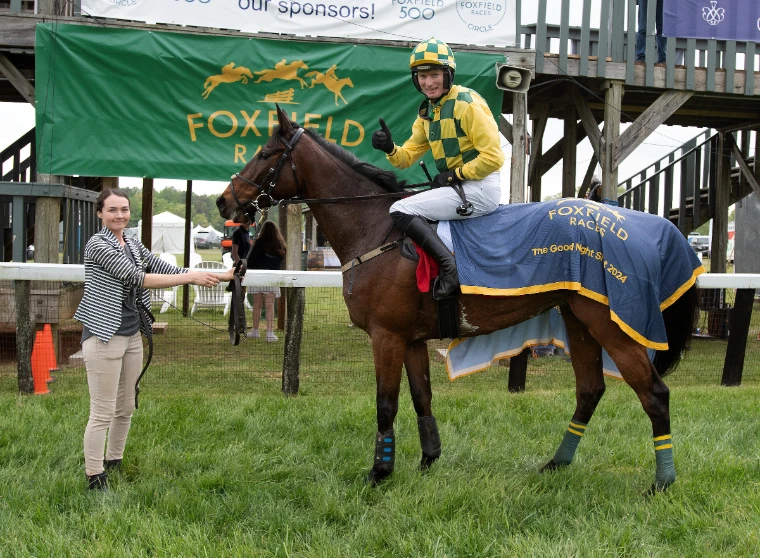 A jockey in green and yellow checks giving a thumbs-up from the saddle of a winning chaser in the enclosure at Foxfield, on the spring timber circuit that leads to The Maryland Hunt Cup, a late-April engagement in the season calendar of The Mansa Court
