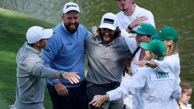 Players and their families in white caddie overalls celebrating at the water during the Par 3 Contest on the Wednesday of The Masters, an engagement in The Mansa Court season