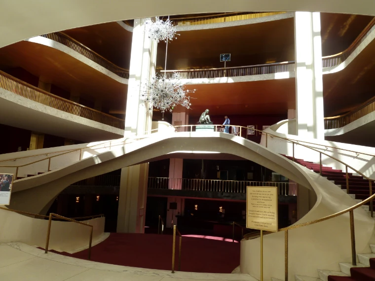 The curving white grand staircase of the Metropolitan Opera lobby with red carpet and brass rails, crystal chandeliers hanging above (2012).