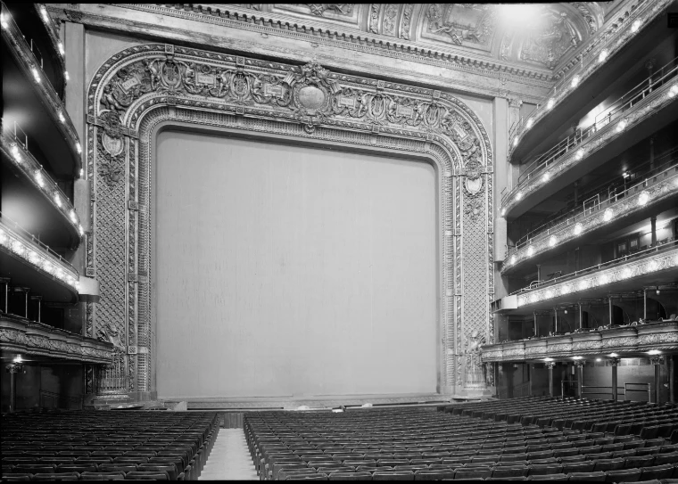 Black-and-white view of the old Metropolitan Opera House auditorium, its ornate gilded proscenium framing the curtain, with tiers of boxes and balconies rising on the right and rows of empty seats below.