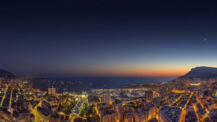Monaco seen from the heights at dusk, its streets and harbour lit gold under a darkening blue sky with a thin crescent moon (2014).