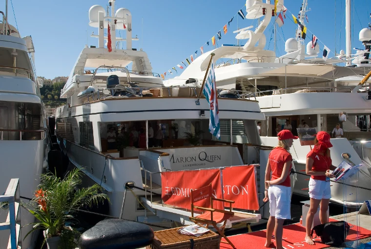 Two crew members in red caps and white trousers stand on a red carpet at the stern of a motor yacht moored stern-to on the show quay, with flag-dressed yachts behind (2011).