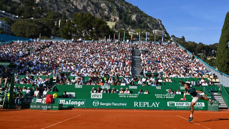 A player serving before a full stand in the evening sun at the Monte-Carlo Country Club, the Rolex Monte-Carlo Masters, an April engagement in the season calendar of The Mansa Court