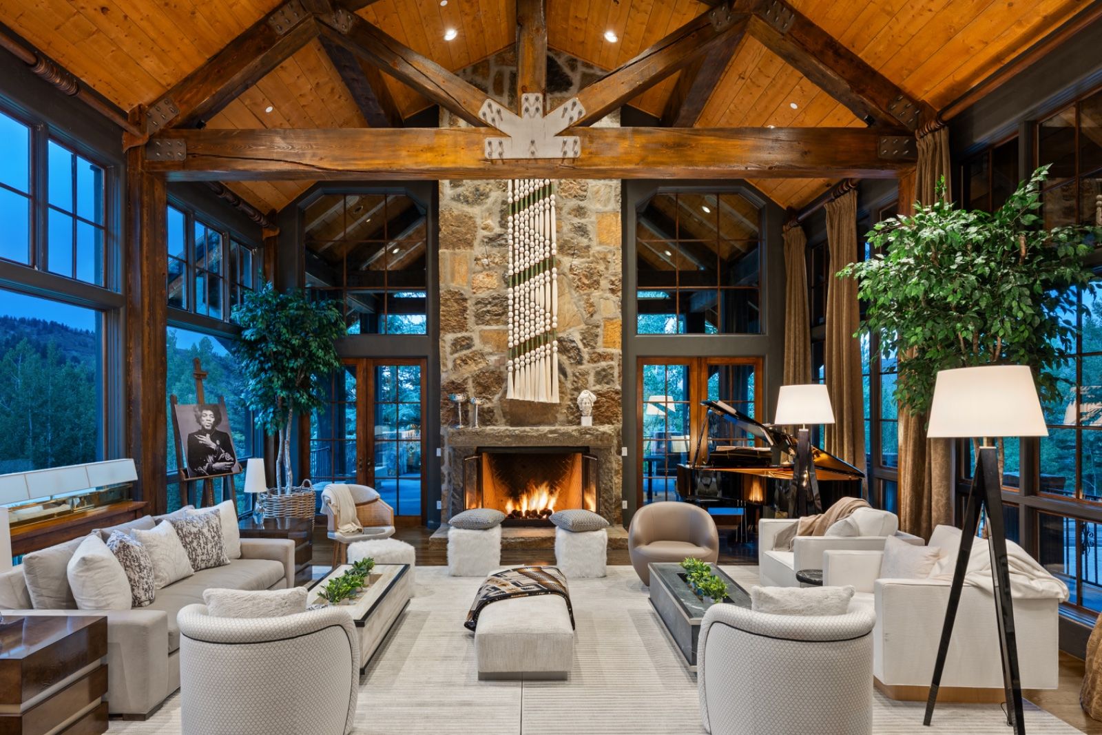 The great room of a timber-framed mountain house at dusk: exposed trusses, a lit stone fireplace, a grand piano, and tall windows onto the aspens