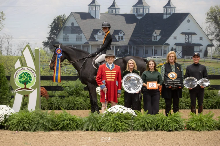 A rider on a dark horse with a championship ribbon, beside a ringmaster in a scarlet coat and presenters holding silver trophies, in front of a painted barn backdrop (2025).