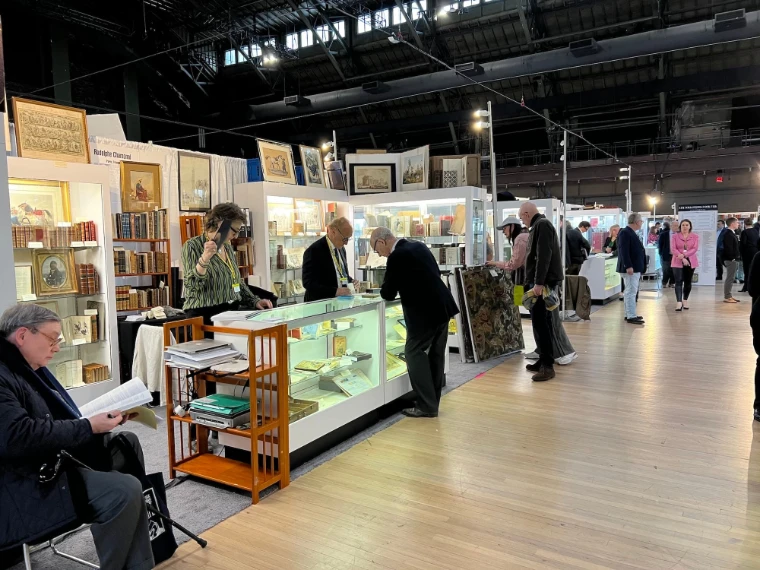 Collectors browse glass cases of rare books and framed prints along an aisle of the fair. The New York International Antiquarian Book Fair, Park Avenue Armory, New York, in the season calendar of The Mansa Court