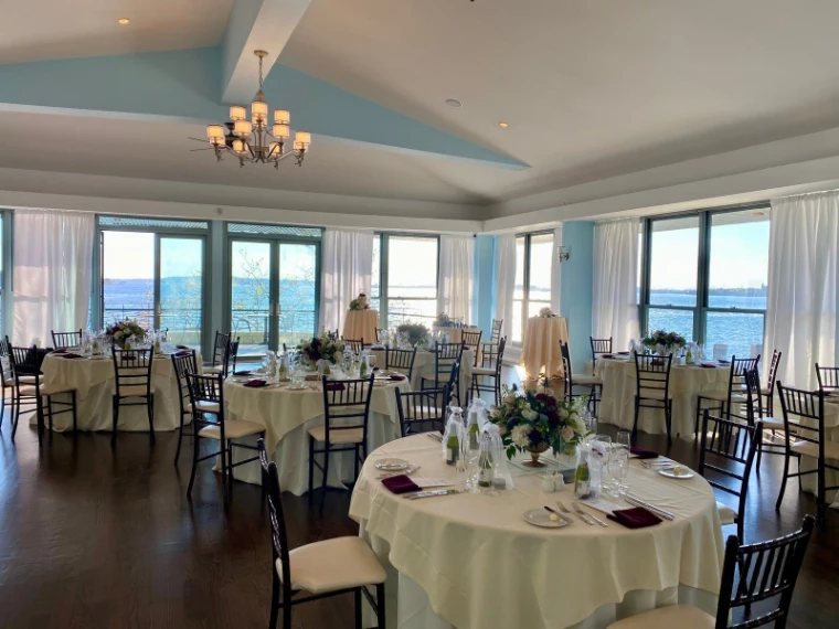 Round tables laid with white cloths, glassware and flowers in a bright room whose windows look out over the water.