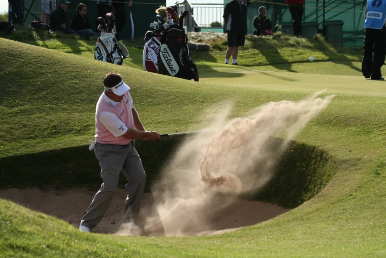 A golfer in a pink sweater blasts a spray of sand out of a deep pot bunker beside the green, with caddies and golf bags on the bank behind (2008).
