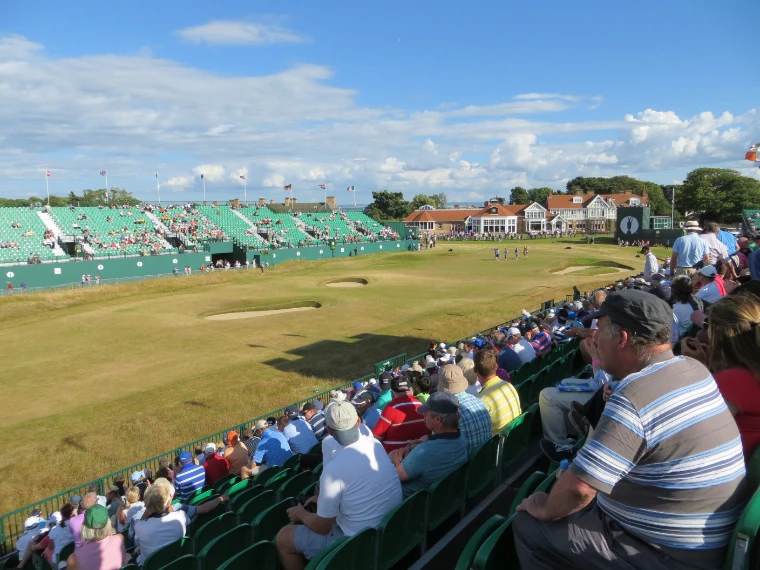 Spectators fill the grandstands along the 18th fairway in long evening light, looking towards the green, more packed stands and the clubhouse (2013).