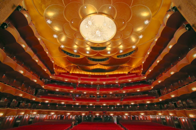 The empty auditorium of the Metropolitan Opera House, its starburst crystal chandeliers lit against the gold-leaf ceiling above tiers of red seats (2016).