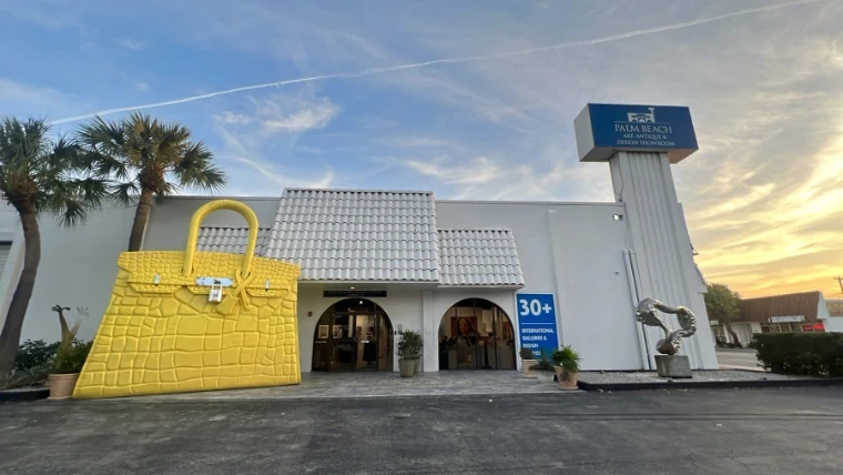 The Palm Beach Art, Antique and Design Showroom at sunset, a giant yellow handbag sculpture beside its arched entrance, an engagement in the season calendar of The Mansa Court