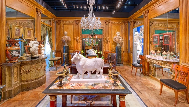 A white marble bull on a specimen-marble table beneath a crystal chandelier, among classical busts and painted urns in a panelled dealer’s stand at a Palm Beach art and antiques fair, an engagement in the season calendar of The Mansa Court
