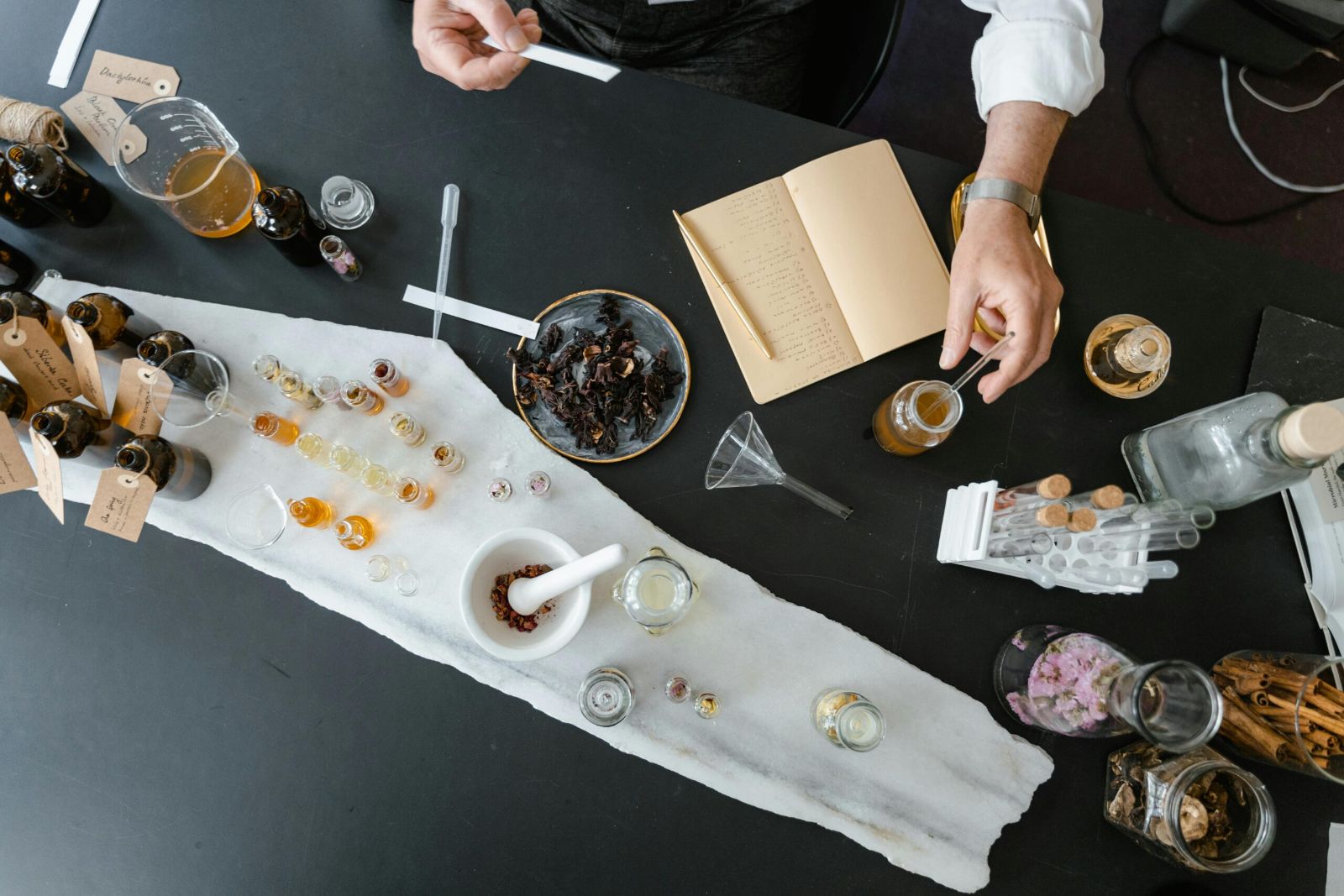A perfumer’s working table seen from above: labelled amber bottles, rows of vials on a marble slab, a mortar of dried petals, a funnel and an open formulation book