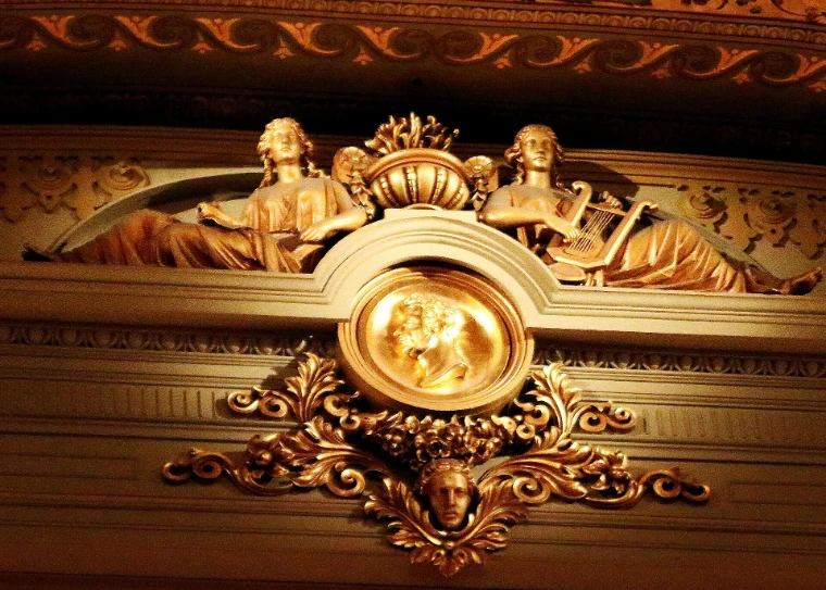 A gilded carving of two reclining figures with a lyre above a medallion, part of the decoration of the Academy of Music’s auditorium (2017).
