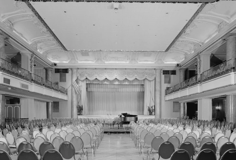A black-and-white view of the second-floor ballroom of the Bellevue-Stratford, rows of chairs facing a curtained stage beneath an ornate plaster ceiling and balconies.