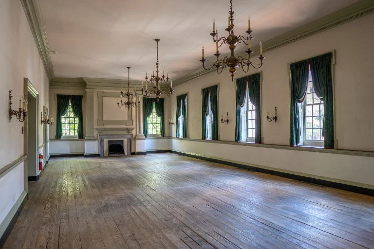 The empty second-floor ballroom of City Tavern, with wide wooden floorboards, brass chandeliers and green-curtained sash windows (2021).