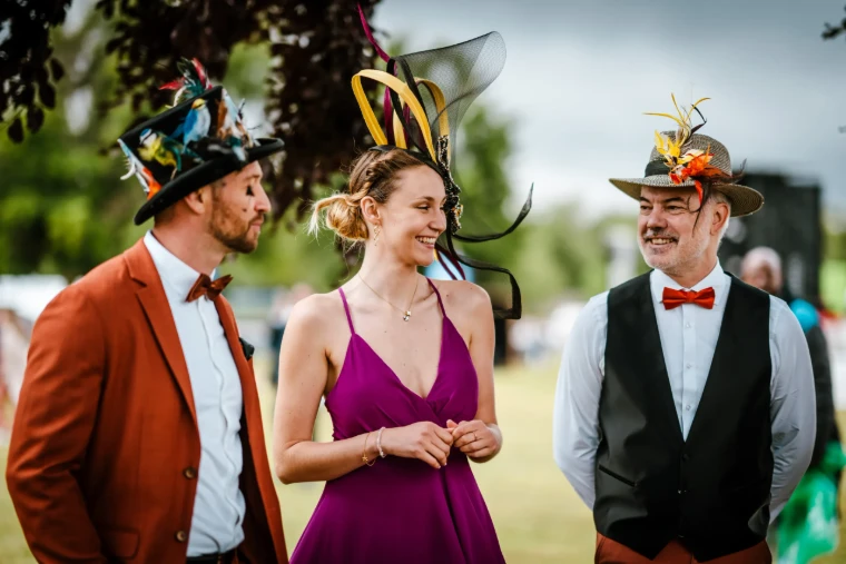 Three racegoers in feathered hats, bow ties and a fuchsia gown talking on the lawn at the Prix de Diane in Chantilly, an engagement in the season calendar of The Mansa Court
